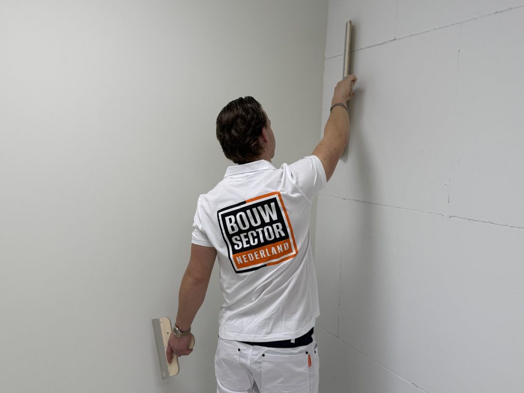 Stukadoor reikt met een smalle spaan naar de bovenkant van een wand in de hoek van een lichte kamer