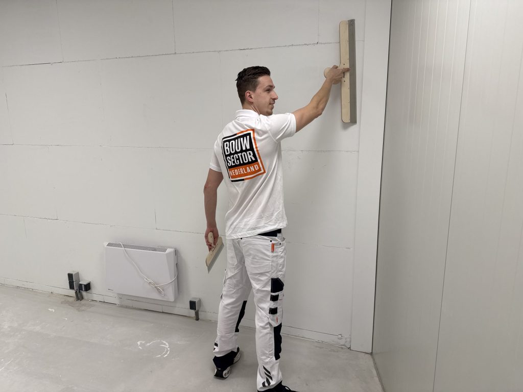 Stukadoor in witte werkbroek kijkt over zijn schouder terwijl hij een spaan tegen een wand boven een kachel houdt