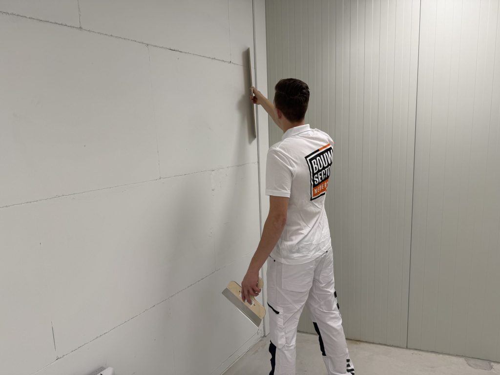 Stukadoor met plakspaan in de hand houdt een lange spaan tegen een wand van grote blokken naast een hoek