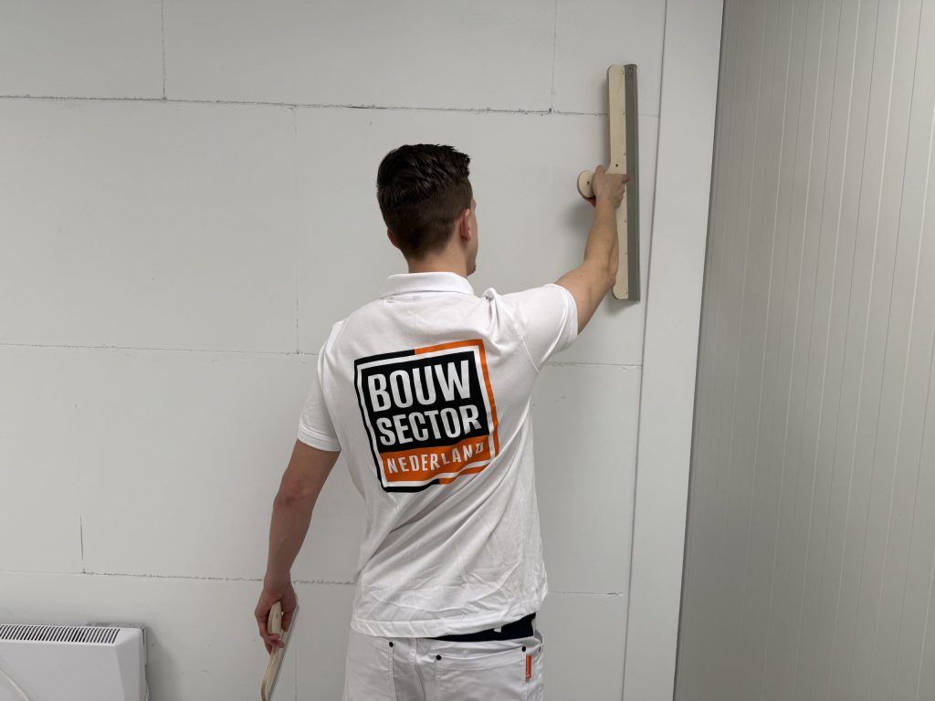 Stukadoor drukt met een hand een houten spaan tegen de bovenkant van een wand van blokken