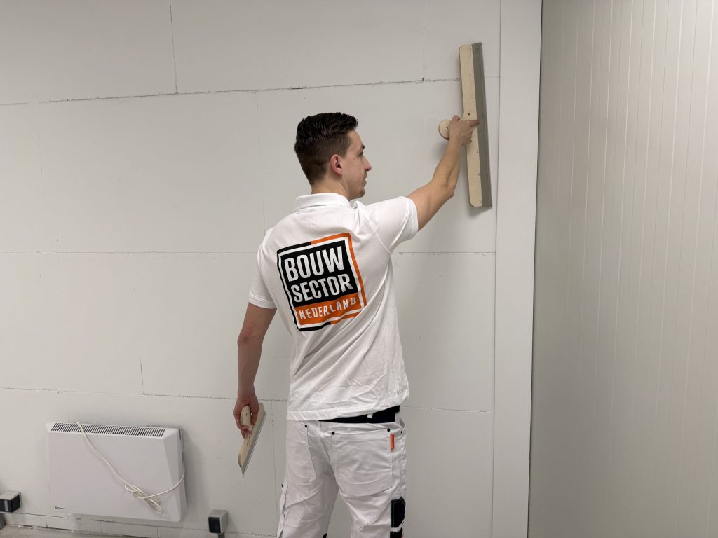 Stukadoor in wit polo met Bouwsector-logo zet een lange spaan hoog tegen een kale binnenwand